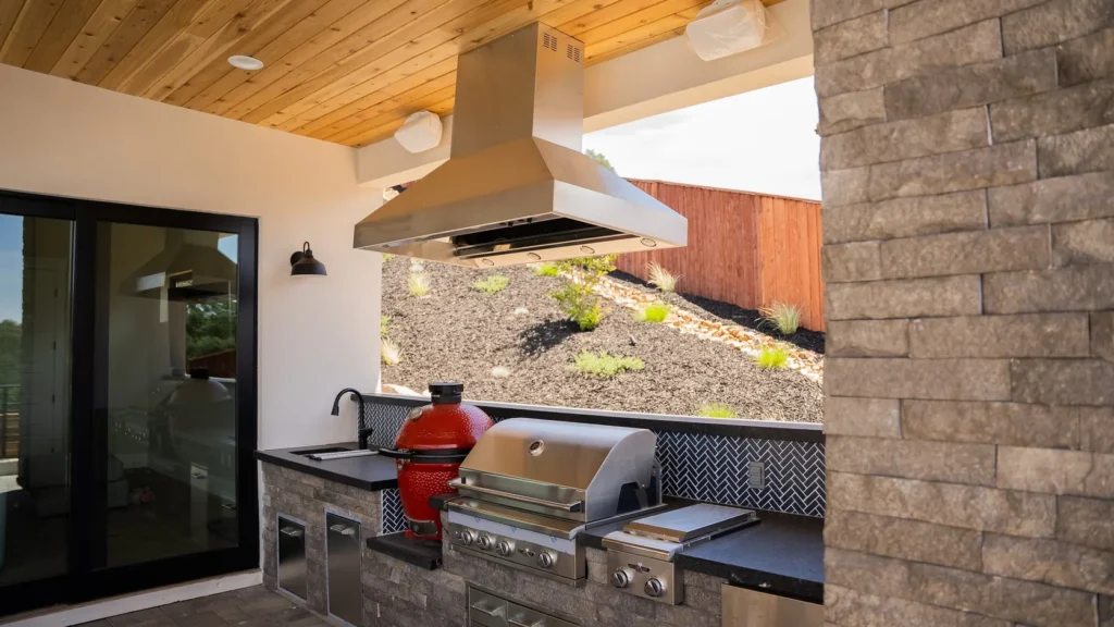 The outdoor kitchen under a cedar-lined ceiling, grill and hood set into stone.